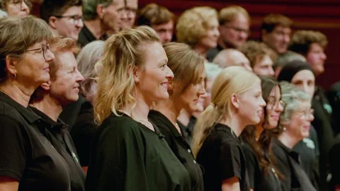 Chor singt auf Bühne, Männer und Frauen in schwarzen Hemden.
