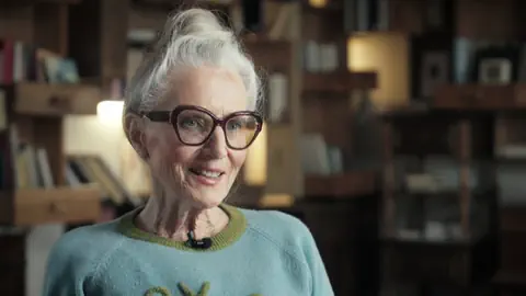 Ältere Frau mit Brille sitzt in einem Raum mit Bücherregalen.