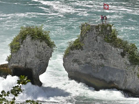 Personen stehen auf der Felsformation mit Schweizer Flagge über rauschendem Wasser.