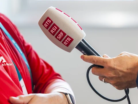 Mikrofon mit Logos von RTS, RSI, SRF, RTR vor rotem Jackett.