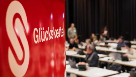 Glückskette-Logo, Menschen bei einer Versammlung im Hintergrund.