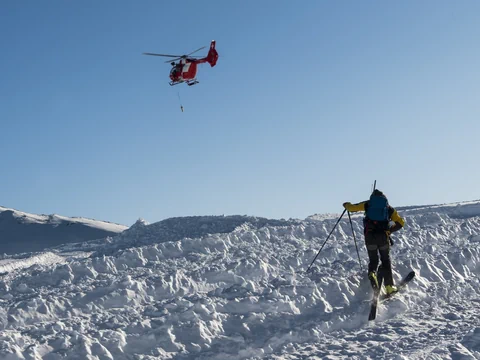 Skifahrer auf verschneitem Hang mit rotem Rega-Helikopter am Himmel.
