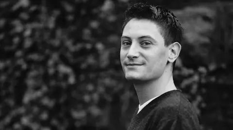 Schwarzweiss Foto von einem jungen Mann, der sich über die Schulter schaut und lächelt.