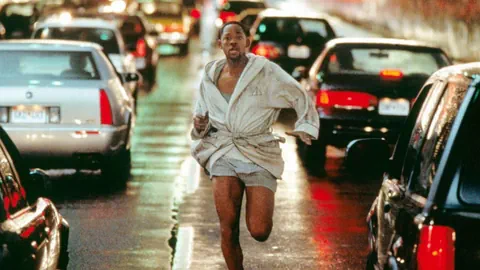 Filmszene: Will Smith rennt in Bademantel und Unterhose in der Mitte einer stark befahrenen Strasse gegen die Verkehrsrichtung.