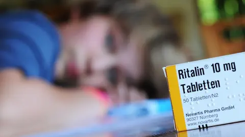 Junge liegt auf einem Tisch und blickt auf eine Packung Ritalin.