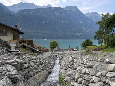 Bach zwischen Felsmauern mit Blick auf See und Berge im Hintergrund.