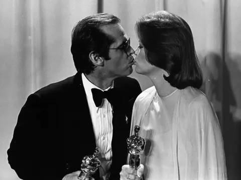 Zwei Personen küssen sich auf einer Preisverleihung, halten Trophäen.