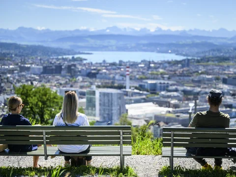 Drei Personen auf Bänken mit Blick auf eine Stadtlandschaft und Berge im Hintergrund.