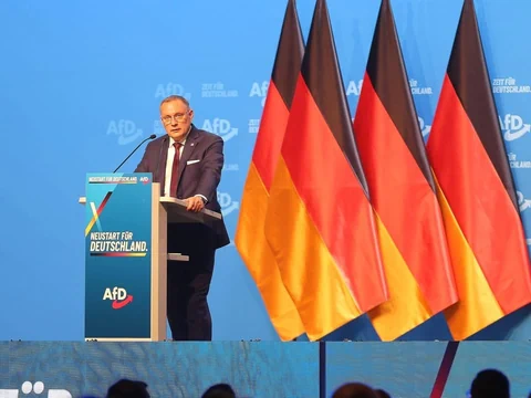 Eine Person steht an einem Rednerpult mit mehreren deutschen Flaggen im Hintergrund.