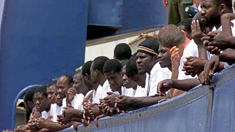 Zahlreiche Afrikaner stehen an einer blauen Schiffsbrüstung.Menschen 