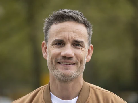 Mann mit brauner Jacke im Freien, unscharfer Hintergrund.