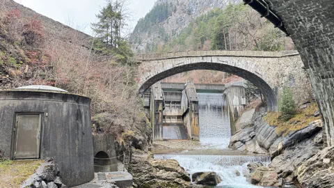 Steinbrücke mit Wasserfall in bewaldeter Berglandschaft.