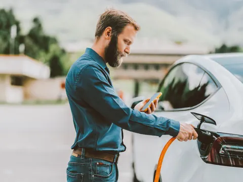 Mann lädt Elektroauto auf und schaut auf sein Handy.