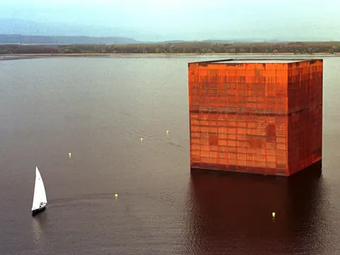 Ein grosses, rostbraunes Würfelgebäude im Wasser neben einem Segelboot.
