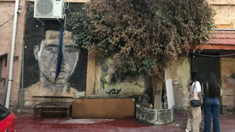 Eine Wand mit einem grossen Porträt und ein Baum daneben; drei Personen stehen davor.