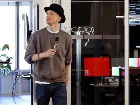 Raphael Labhart kommt in ein Medienbüro mit Detektivhut und einer Lupe in der Hand.