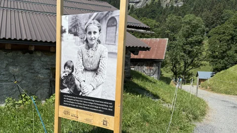 Ein grosses Holzschild mit einem schwarz-weissen Foto und Text steht neben einem ländlichen Weg und Gebäuden, umgeben von Bäumen und Wiesen.
