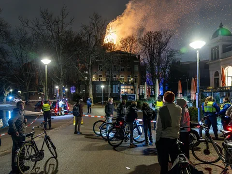 Leute mit Velos warten an einer Polizeiabsperrung. Im Hintergrund der brennende Kirchturm. 