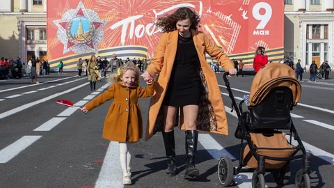 Frau mit Kind und Kinderwagen auf Strasse vor Plakat.