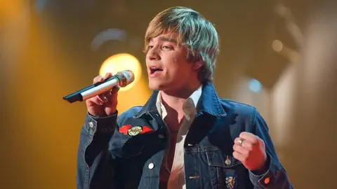 Ein junger Mann mit blonden Haaren und Jeansjacke singt in ein Mikrophon