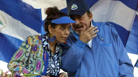 Mann und Frau stehen vor wehenden Flaggen, eine trägt eine blaue Kappe und bunte Kleidung.