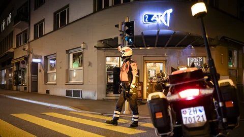 Nachtszene mit einer Person in Motorradkleidung vor einem Gebäude und einem geparkten Motorrad auf der Strasse.