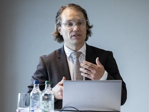 Mann im Anzug spricht auf einer Konferenz neben Laptop und Wasserflaschen.