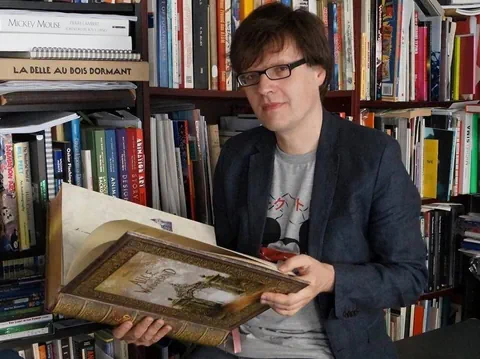 Mann mit Brille sitzt vor einem Bücherregal und schlägt ein übergrosses und dickes Buch auf
