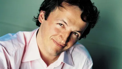 Der Dirigent Jean-Christophe Spinosi in rosa Hemd vor grünem Hintergrund. 