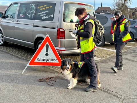 Zwei Personen in gelben Westen mit einem Hund neben einem Warnschild im Freien.