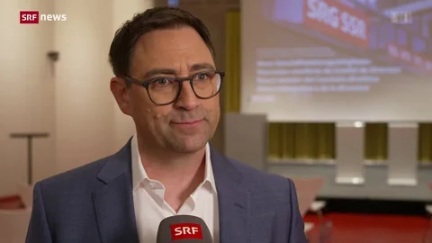 Person mit Brille und Anzug hält ein SRF-Mikrofon, verschwommener Hintergrund mit Leinwand.