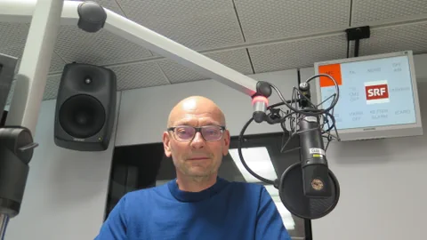 Ein Mann mit Glatze und Brille hinter einem Mikrofon.