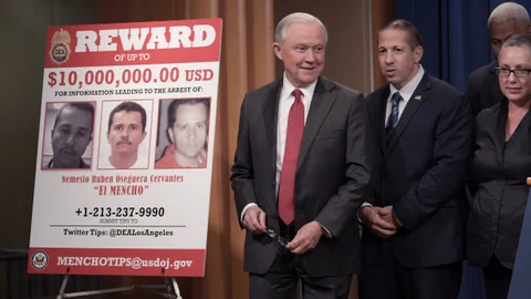 Mann in Anzug spricht neben Belohnungsplakat mit Fotos gesuchter Personen.