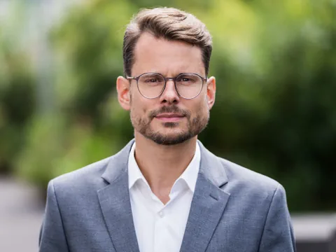 Mann in Anzug mit Brille im Freien vor unscharfem Hintergrund.