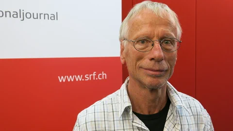 Mann mit grauen Haaren und Brille lächelt leicht in die Kamera. Er steht vor einem roten Hintergrund.