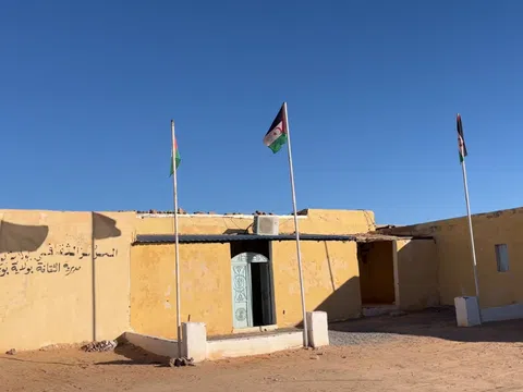 Gelbes Gebäude mit drei Fahnenmasten in Wüstenlandschaft.