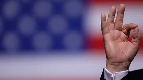 Hand zeigt OK-Geste vor US-Flagge.