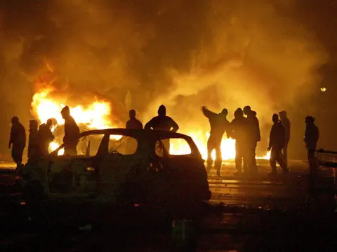 Menschen vor brennendem Auto und Flammen in der Nacht.