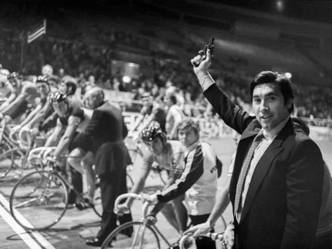 Eine Person hebt eine Pistole in die Luft vor Radfahrern auf der Startlinie in einem Stadion.