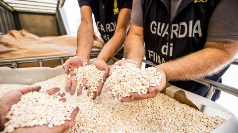 Personen halten weisse Tabletten in den Händen, hinter ihnen steht 'Guardia di Finanza' auf Uniformen geschrieben.