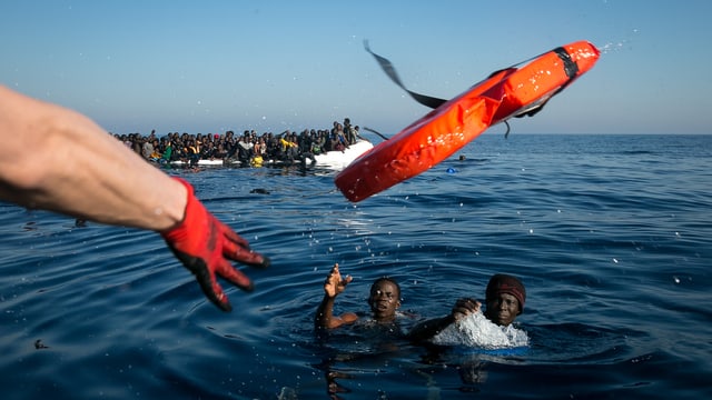 Ein Arm mit rotem Handschuh wirft zwei im Meer treibenden Flüchtlingen eine Schwimmweste zu. Ein Arm mit rotem Handschuh wirft zwei im Meer treibenden Flüchtlingen eine Schwimmweste zu.