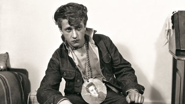 Mann sitzt auf Sessel mit Elvis-Plakette um den Hals Mann sitzt auf Sessel mit Elvis-Plakette um den Hals