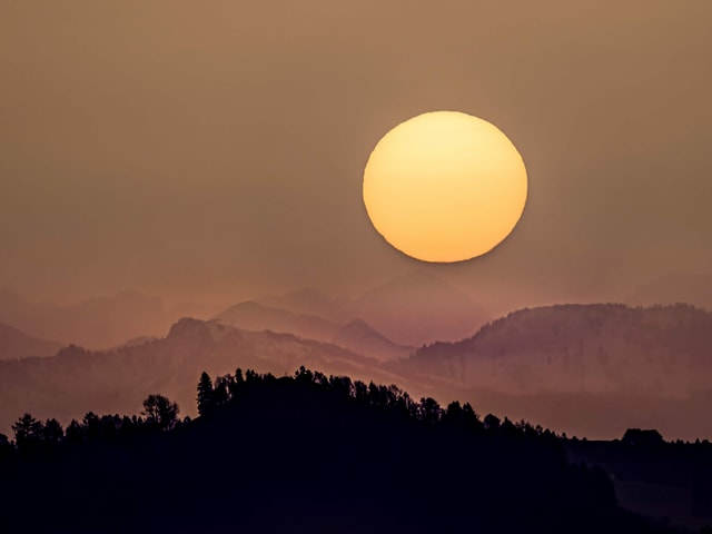 Bräunlicher Himmel mit gelber Sonnenscheibe. Im Vordergrund dunkle Konturen von Hügeln.