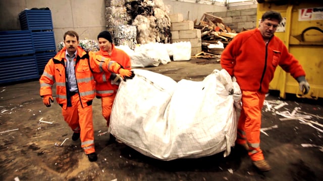 Drei Männer in orangen Uniformen tragen einen riesigen Müllsack. Drei Männer in orangen Uniformen tragen einen riesigen Müllsack.