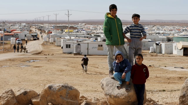 Im Vordergrund vier Kinder, im Hintergrund in wüstenähnlichem Gebiet ein Flüchtlingscamp aus Containern. Im Vordergrund vier Kinder, im Hintergrund in wüstenähnlichem Gebiet ein Flüchtlingscamp aus Containern.