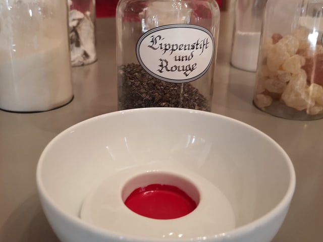 Weisses Schälchen mit roter Paste. Dahinter Glas mit Aufschrift "Lippenstift und Rouge". Weisses Schälchen mit roter Paste. Dahinter Glas mit Aufschrift "Lippenstift und Rouge".