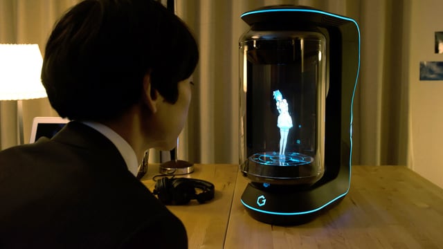 Ein junger Mann im Anzug sitzt am Tisch uns spricht zu einem Hologramm hinter der Glasscheibe. Ein junger Mann im Anzug sitzt am Tisch uns spricht zu einem Hologramm hinter der Glasscheibe.