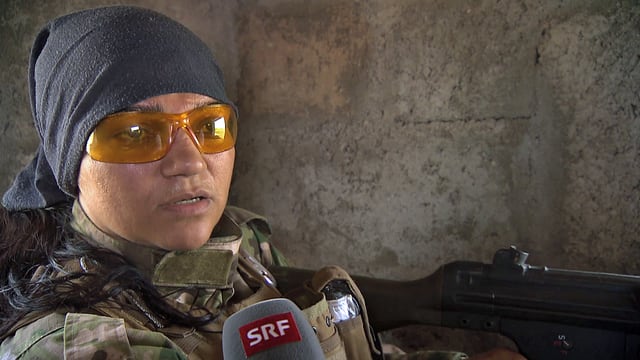 Eine Frau mit Kopftuch und Sonnenbrille mit einem Gewehr im Gespräch mit SRF. Eine Frau mit Kopftuch und Sonnenbrille mit einem Gewehr im Gespräch mit SRF.