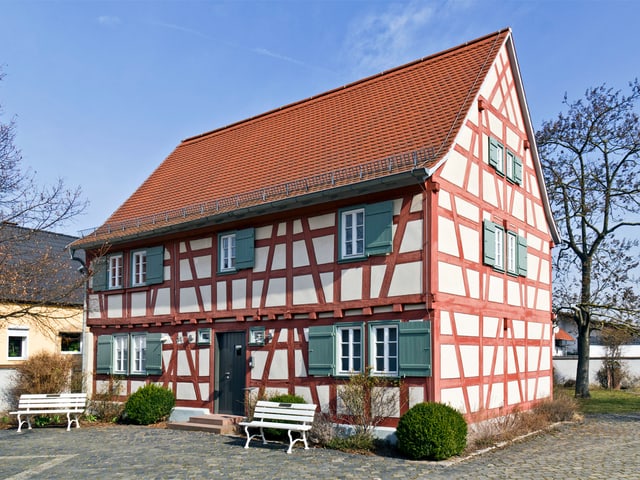Ein Riegelhaus mit grünen Fensterläden, davor stehen zwei weisse Sitzbänke und ein paar Büsche. Ein Riegelhaus mit grünen Fensterläden, davor stehen zwei weisse Sitzbänke und ein paar Büsche.