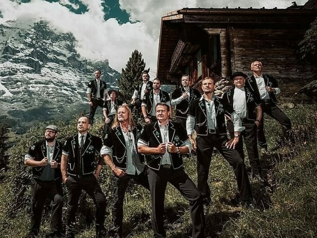 Männercombo steht vor Alphütte an Abhang in Bergkulisse. Männercombo steht vor Alphütte an Abhang in Bergkulisse.